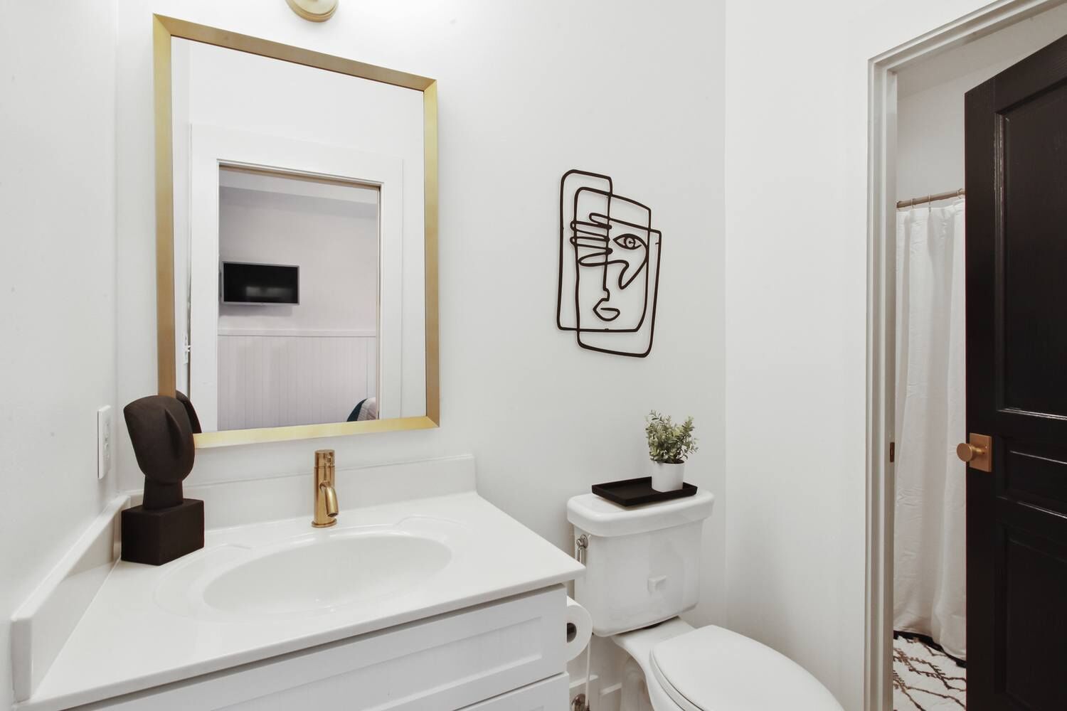 Une salle de bain moderne avec un lavabo blanc, un robinet doré et un grand miroir avec un cadre doré. On y trouve des toilettes sur lesquelles est posée une petite plante, ainsi qu’une œuvre d’art abstraite en noir et blanc accrochée au mur.