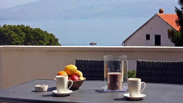 Une table avec un panier de fruits, deux tasses et une bougie, sur un balcon donnant sur un lac, avec des montagnes en arrière-plan.