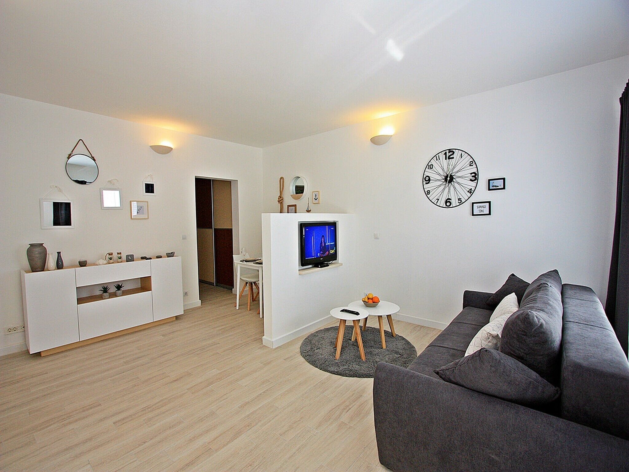 A modern living room with a grey sofa, a TV, a dining area with a white table, and a kitchen area with a white cabinet.