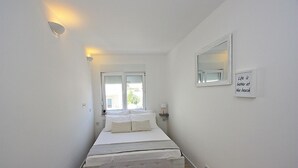 Une chambre étroite aux murs blancs, avec un lit, un banc et un miroir accroché au mur.