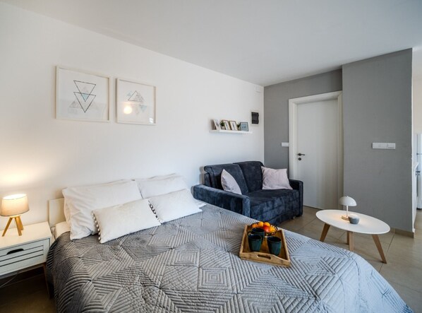 A bedroom with a bed, a bedside table, a chair, a small table with a tray of fruit, and wall art.