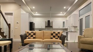 A modern living room with a glass coffee table, a sofa with checkered cushions, and a kitchen in the background. - Dazzling Home Next to Beach & Go Station (Pickering)