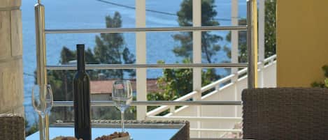A dining table with a bottle and glasses, wicker chairs, and a view of the sea through a window.
