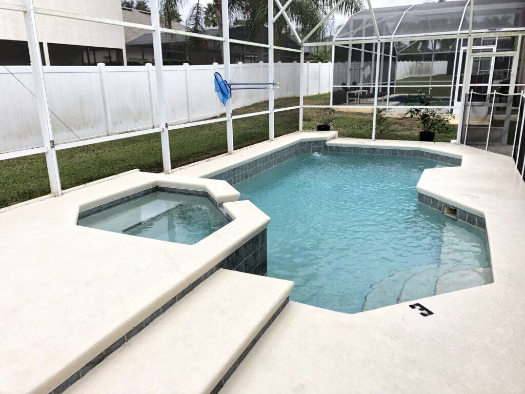 A swimming pool with a hot tub, surrounded by a white fence and a greenhouse structure.