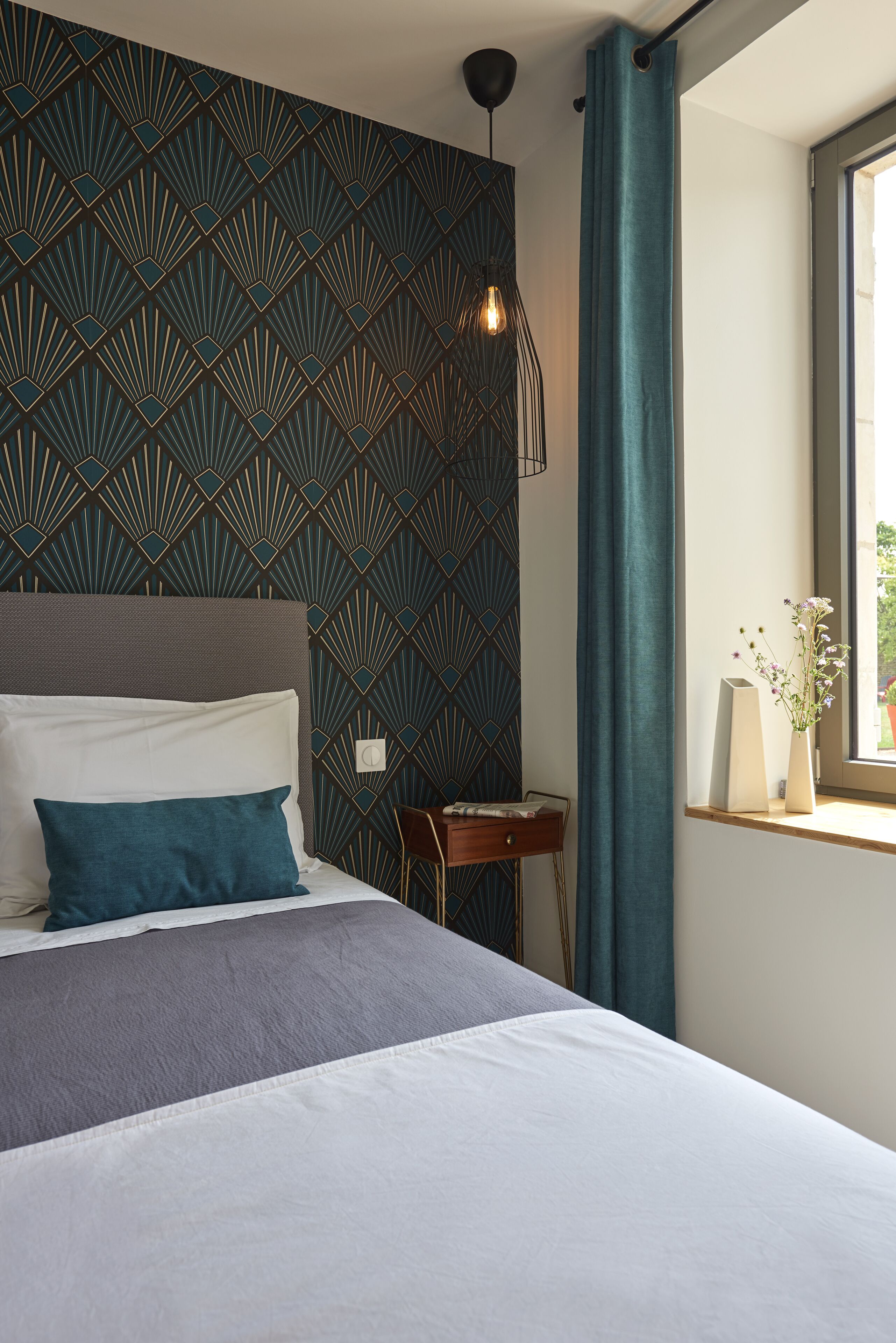 A modern hotel room with a geometric patterned wall, a bed with white and teal bedding, a bedside table with a vase of flowers, and a window with a view of greenery.