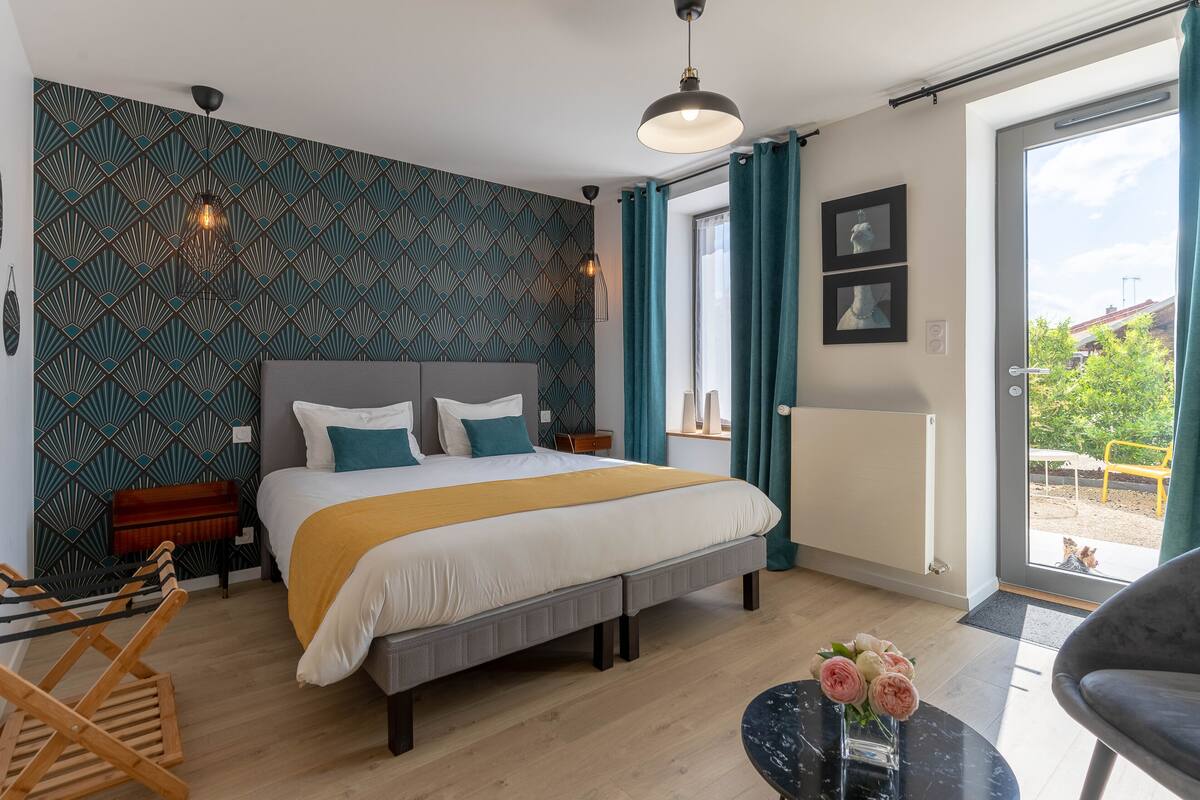 Ein modernes Schlafzimmer mit einem großen Bett, einem Holzstuhl, einem kleinen Tisch mit Blumen und Blick ins Freie durch eine Glastür.