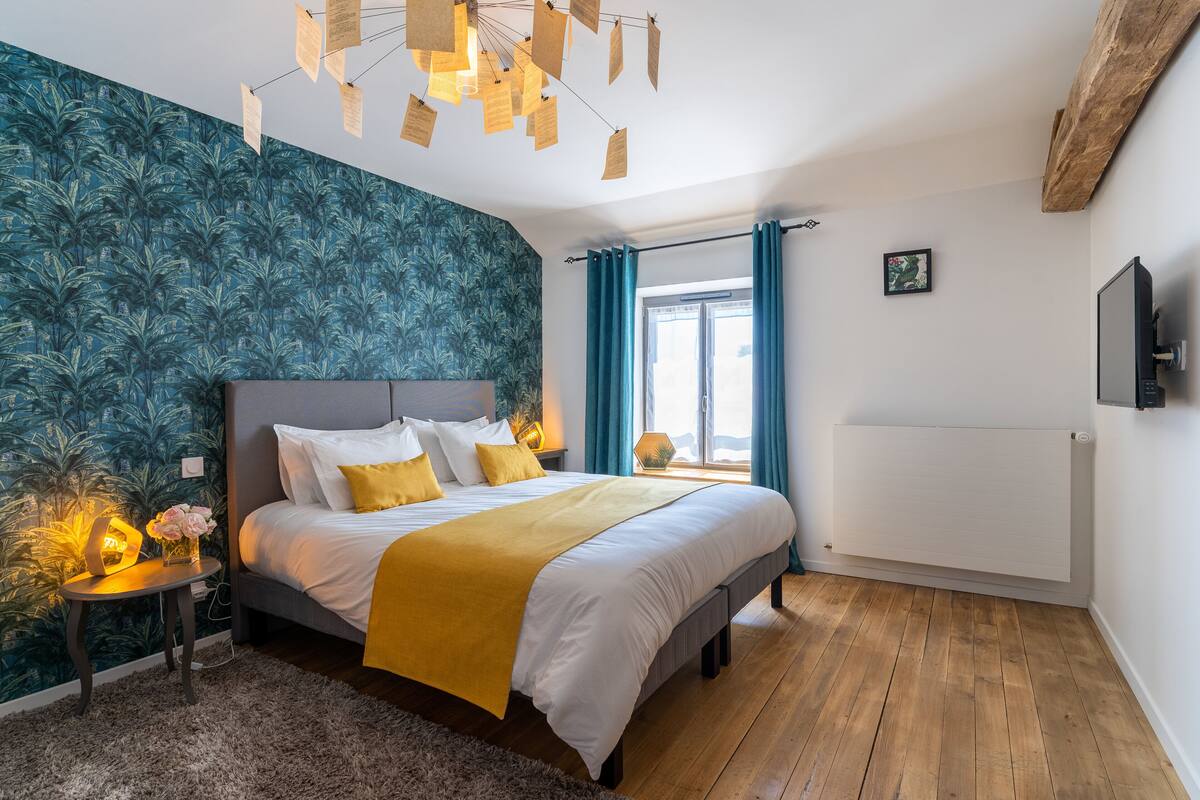 Ein Schlafzimmer mit Bett, Nachttisch, Wand-Fernseher, Fenster mit Vorhängen und Heizkörper.