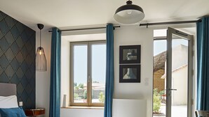 Ein Schlafzimmer mit Bett, Nachttisch, Kommode, Fenster mit Vorhängen und Blick nach draußen.