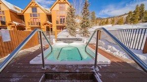 Une cabane en bois avec un bain à remous à l’avant, entourée de neige et d’arbres.