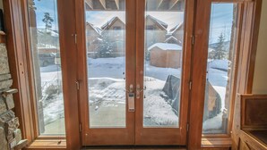 Une porte à deux battants en bois, avec des panneaux de verre, qui donne sur un espace extérieur enneigé où se trouvent des maisons et où le ciel est dégagé.