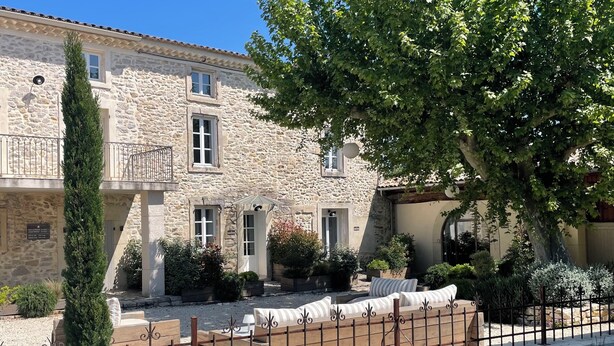 Un edificio de piedra con balcón, mesas al aire libre y un árbol en primer plano.