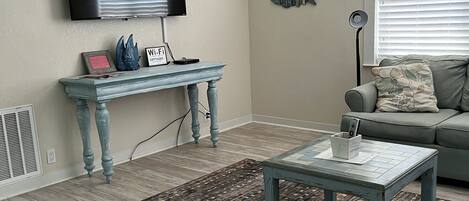 A living room with a wooden floor, a blue coffee table, a grey sofa, a TV mounted on the wall, and a ceiling fan.