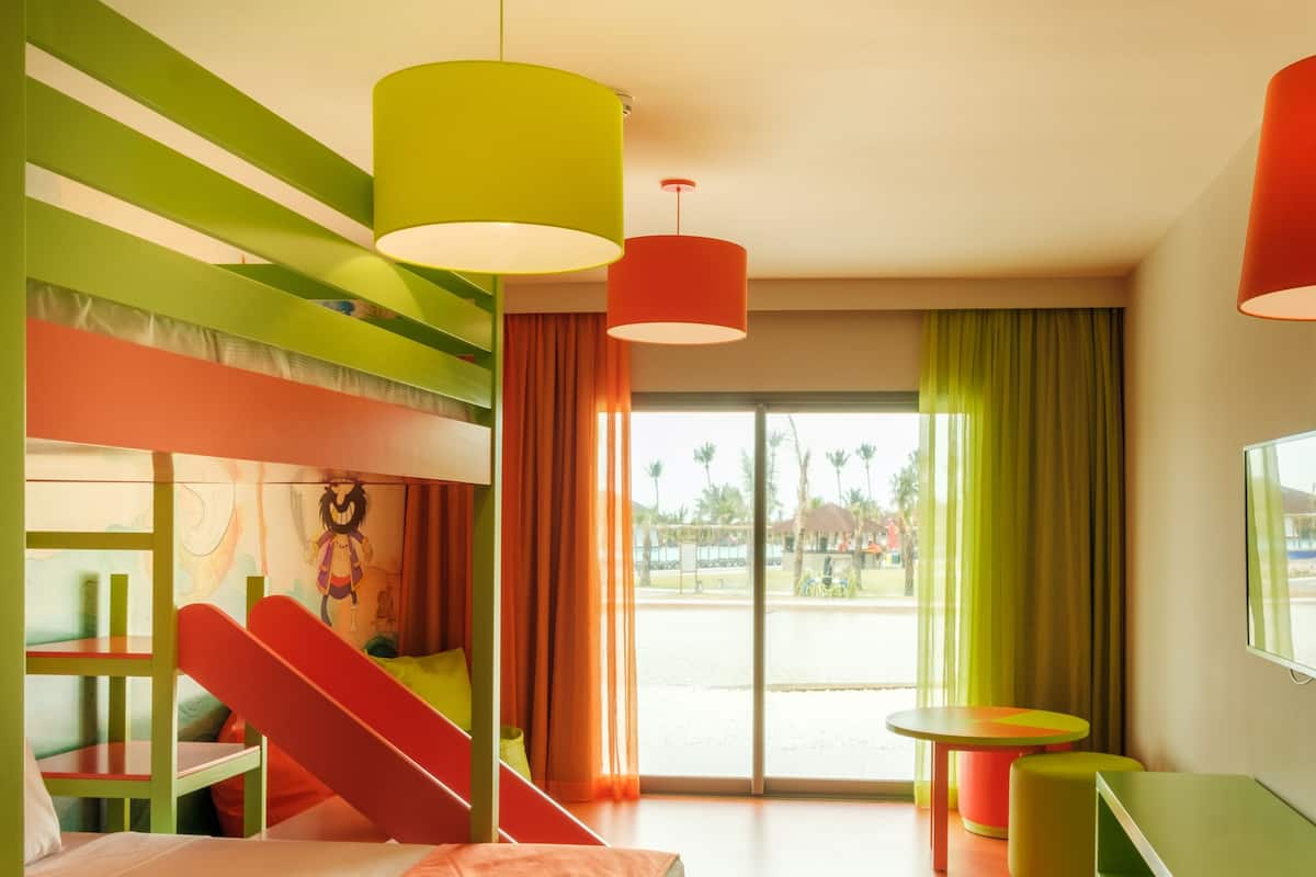 Una habitación luminosa y colorida con literas, una mesita pequeña y una ventana con cortinas.