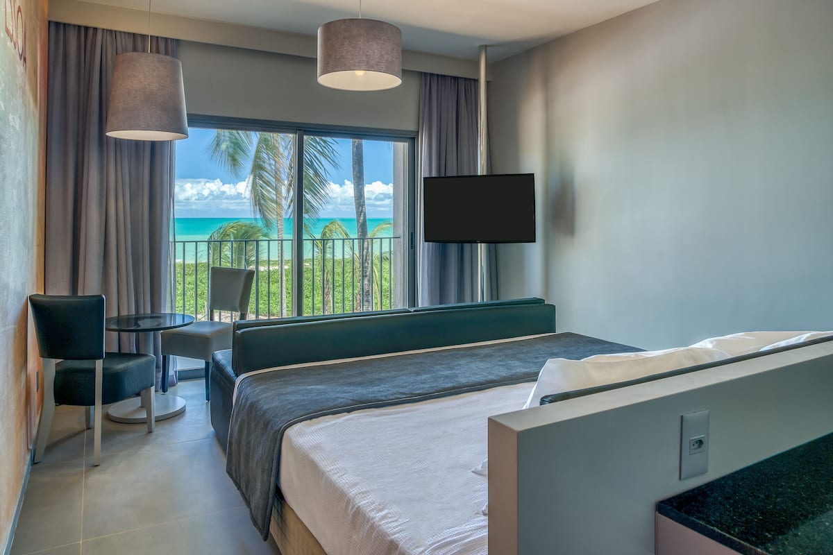 Habitación de hotel con cama, televisor, una mesita pequeña y una silla. Tiene un balcón con vistas al océano y palmeras.