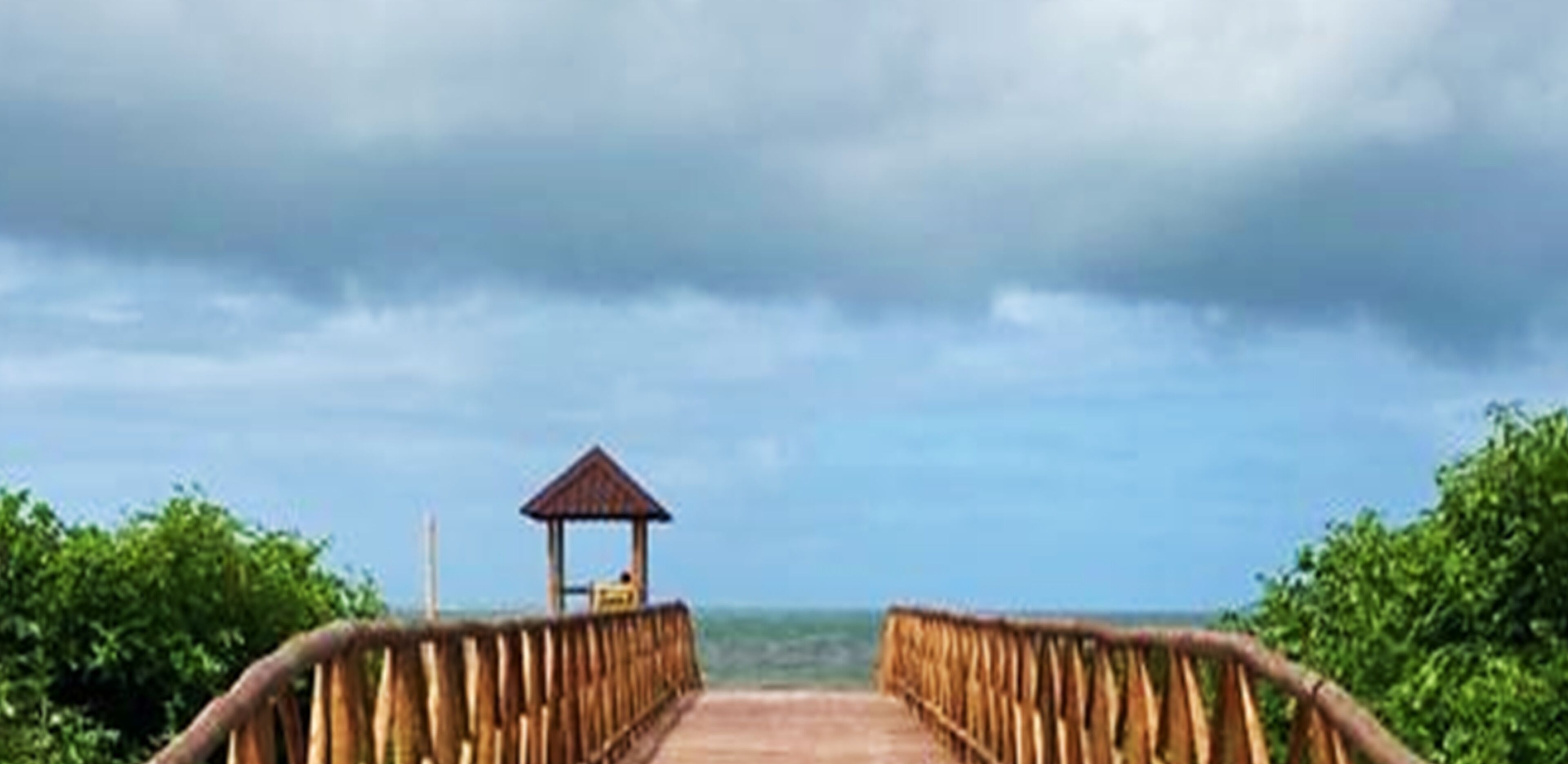 A wooden pier leading to a small hut with a thatched roof, surrounded by lush greenery.