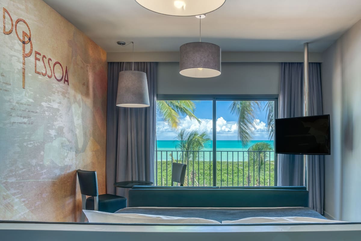 Una habitación de hotel moderna con un amplio ventanal que ofrece vista al océano, una cama, un televisor montado en la pared y una zona de estar con una silla.