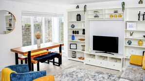 Ein modernes Wohnzimmer mit einem Holzesstisch, zwei Stühlen, einem Sofa und einem Fernseher auf einer weißen Unterhaltungsecke.