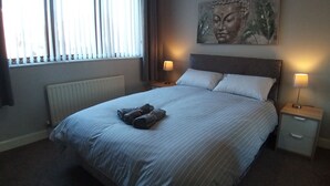 Een opgemaakt bed met kussens en een gevouwen handdoek, twee bedlampen, een radiator en een boeddha-schilderij aan de muur.