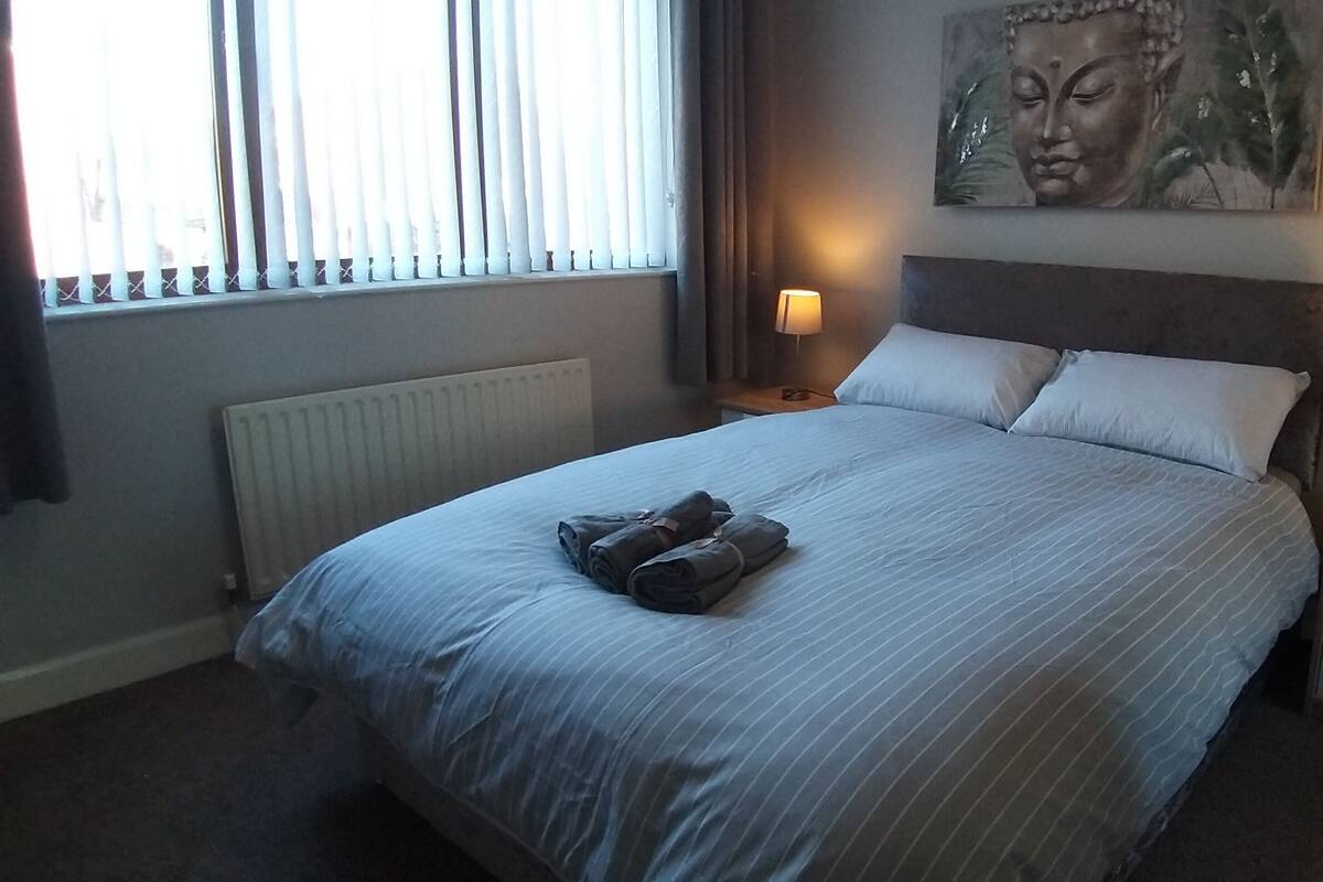 A neatly made bed with pillows and a folded towel, two bedside lamps, a radiator, and a Buddha painting on the wall.