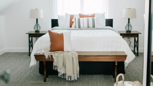 Un dormitorio con una cama grande, dos mesitas de noche con lámparas, un banco y una ventana con cortinas.