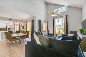 A modern living room with a sofa, dining table, and chairs. There is a ceiling fan, large windows, and a kitchen in the background. - beautiful townhouse in Isla Del Mar (St. Petersburg)