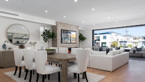 A modern living room with a dining area, white furniture, a fireplace, and a large window with a city view. - Brand New Luxury Beach Home (Newport Beach)