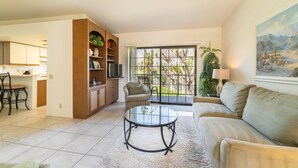 Un soggiorno con un tavolino in vetro, una poltrona, un'area pranzo con una sedia, un angolo cottura con mobili e vista su un giardino tramite una porta scorrevole.