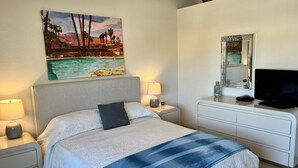 Una camera d'hotel con un letto, un comodino con una lampada, una cassettiera con televisore e un quadro raffigurante una spiaggia.
