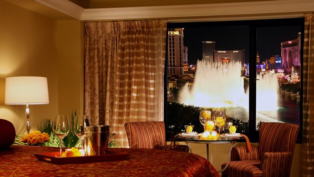 A hotel room with a bed, a dining table with chairs, a view of a cityscape and a fountain.