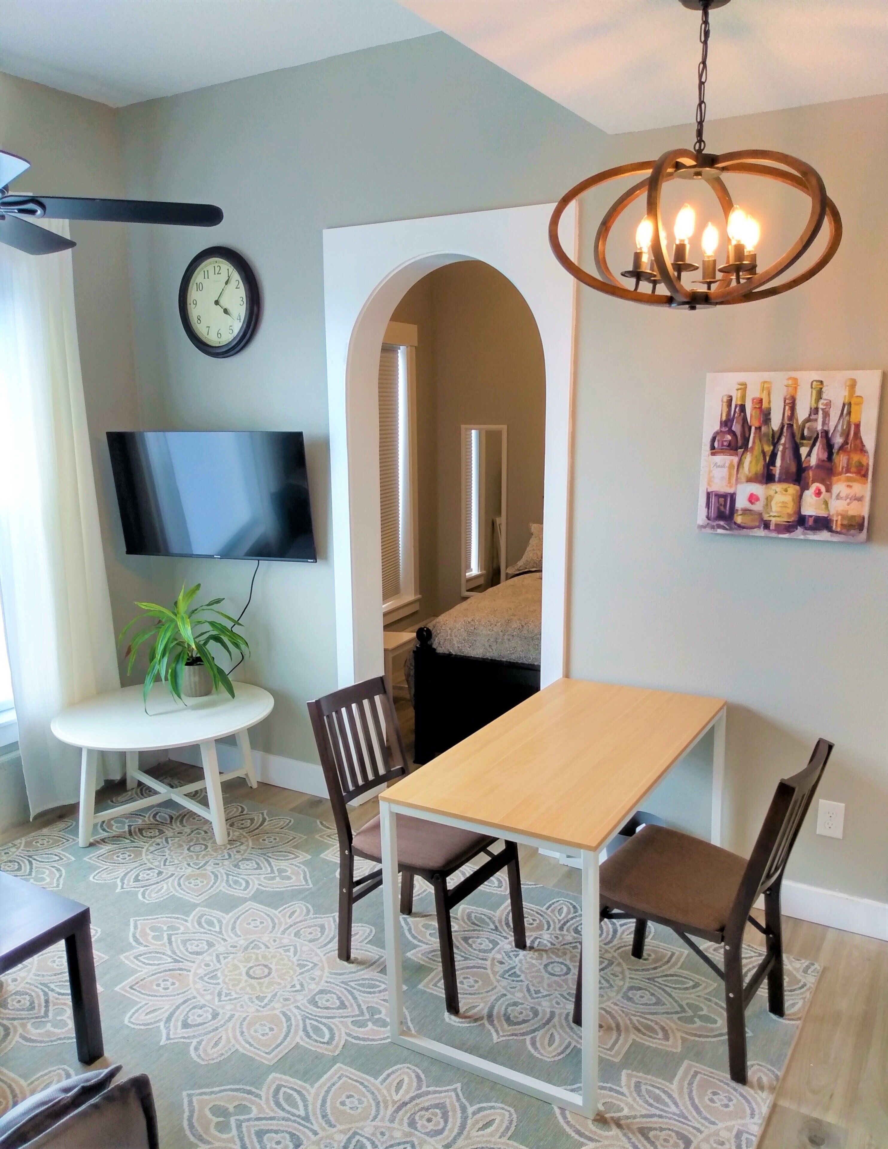 A dining area with a wooden table and chairs, a potted plant, a wall-mounted TV, a clock, and a wine bottle painting.