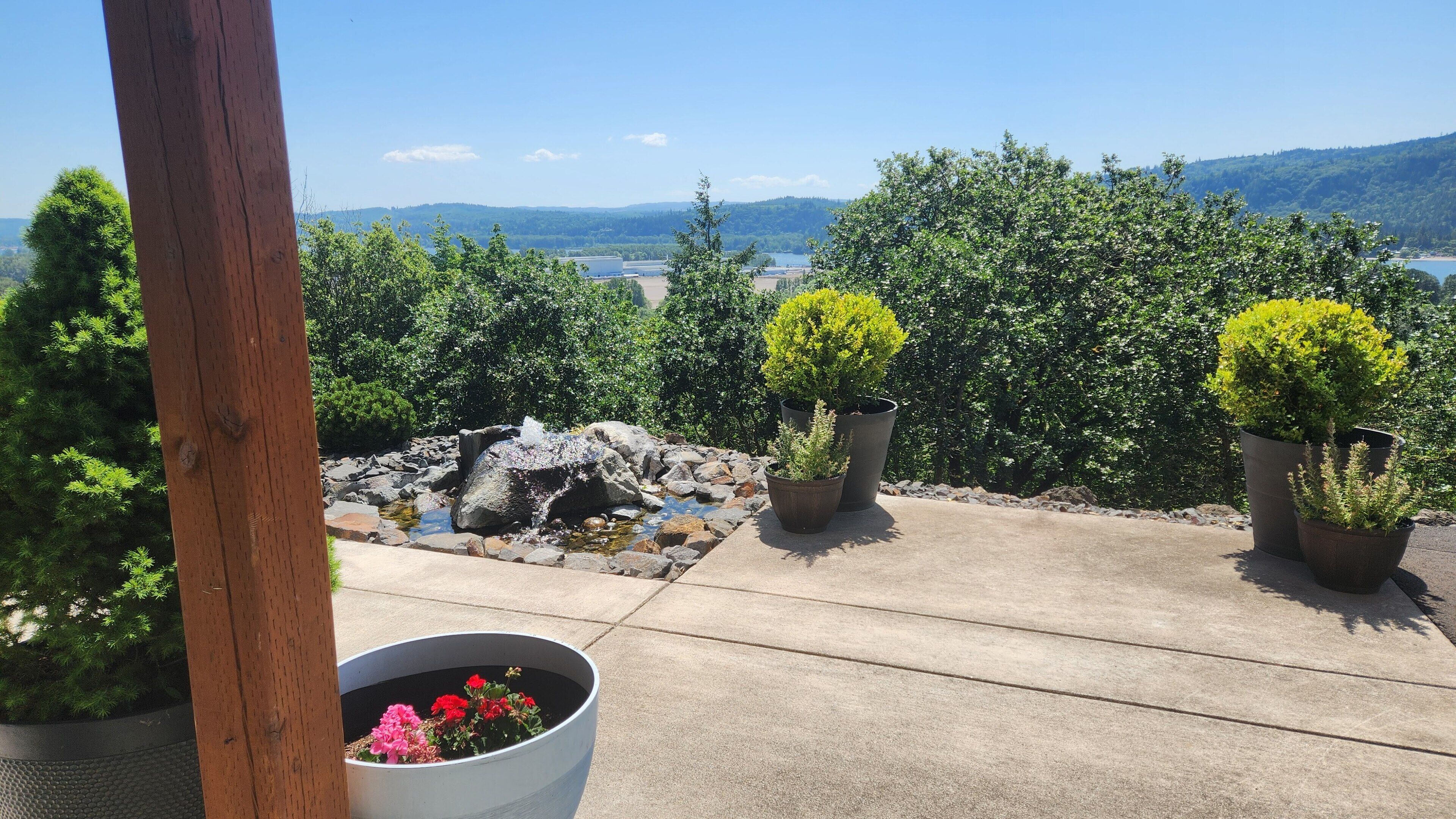 A rooftop garden with potted plants, a small water feature, and a view of a lake and mountains.
