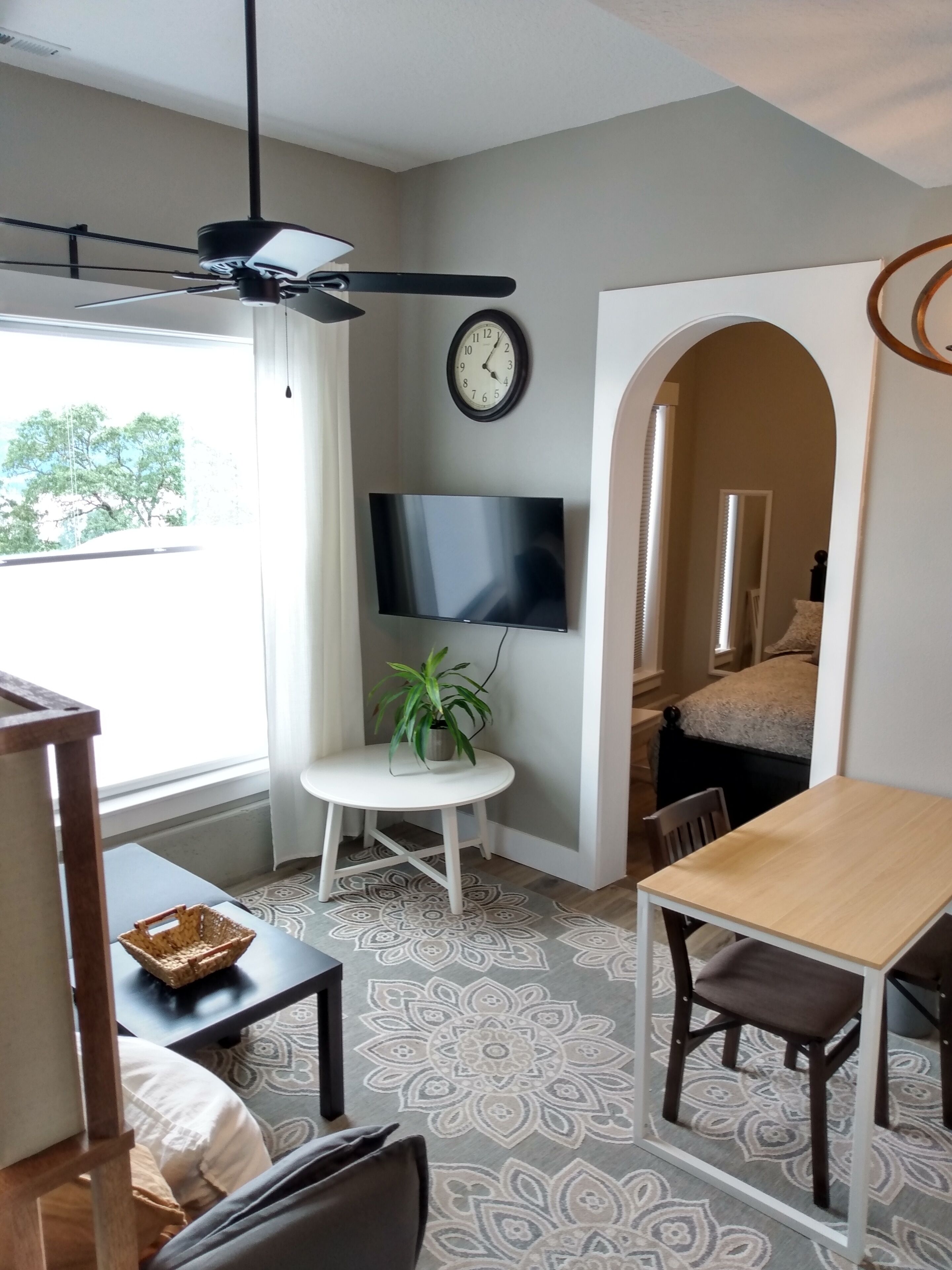 Un salon moderne doté d’un ventilateur de plafond, d’un téléviseur à écran plat, d’une horloge et d’un coin repas avec une table et des chaises.