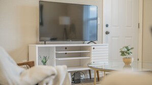 Una sala de estar con un televisor de pantalla plana sobre un soporte blanco, una mesa de centro de vidrio, un sofá blanco y una pequeña planta en maceta.