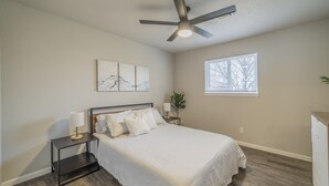 Un dormitorio con cama, mesita de noche, ventilador de techo, ventana y obra de arte en la pared.