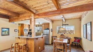 A kitchen with wooden beams, a central island with stools, and a dining area with chairs and a table. - Fort Marcy 57 | Walk to Plaza | Kiva Fireplace | Full Kitchen | Hot Tub | Pool | Single Level (Santa Fe)