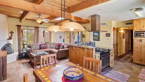 A cozy living room with a fireplace, wooden ceiling beams, a dining table with a colorful table runner, and a kitchen area with wooden cabinets and a stove. - Fort Marcy 57 | Walk to Plaza | Kiva Fireplace | Full Kitchen | Hot Tub | Pool | Single Level (Santa Fe)