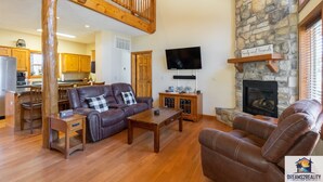 A spacious living room with a stone fireplace, wooden furniture, and a kitchen in the background. - 6BR Branson Lodge | 6 King Rooms | Near Silver Dollar City + Pools (FHL-263WO) (Branson)