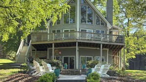 A two-story house with a balcony, a chimney, and a front porch with chairs. - Relaxing Lakefront A-Frame with Hot tub and Firepit on Lake Gaston! (Boydton)