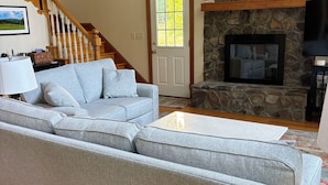 A living room with a grey sectional sofa, a stone fireplace, a large clock, and a wooden staircase. - Relaxing Lakefront A-Frame with Hot tub and Firepit on Lake Gaston! (Boydton)