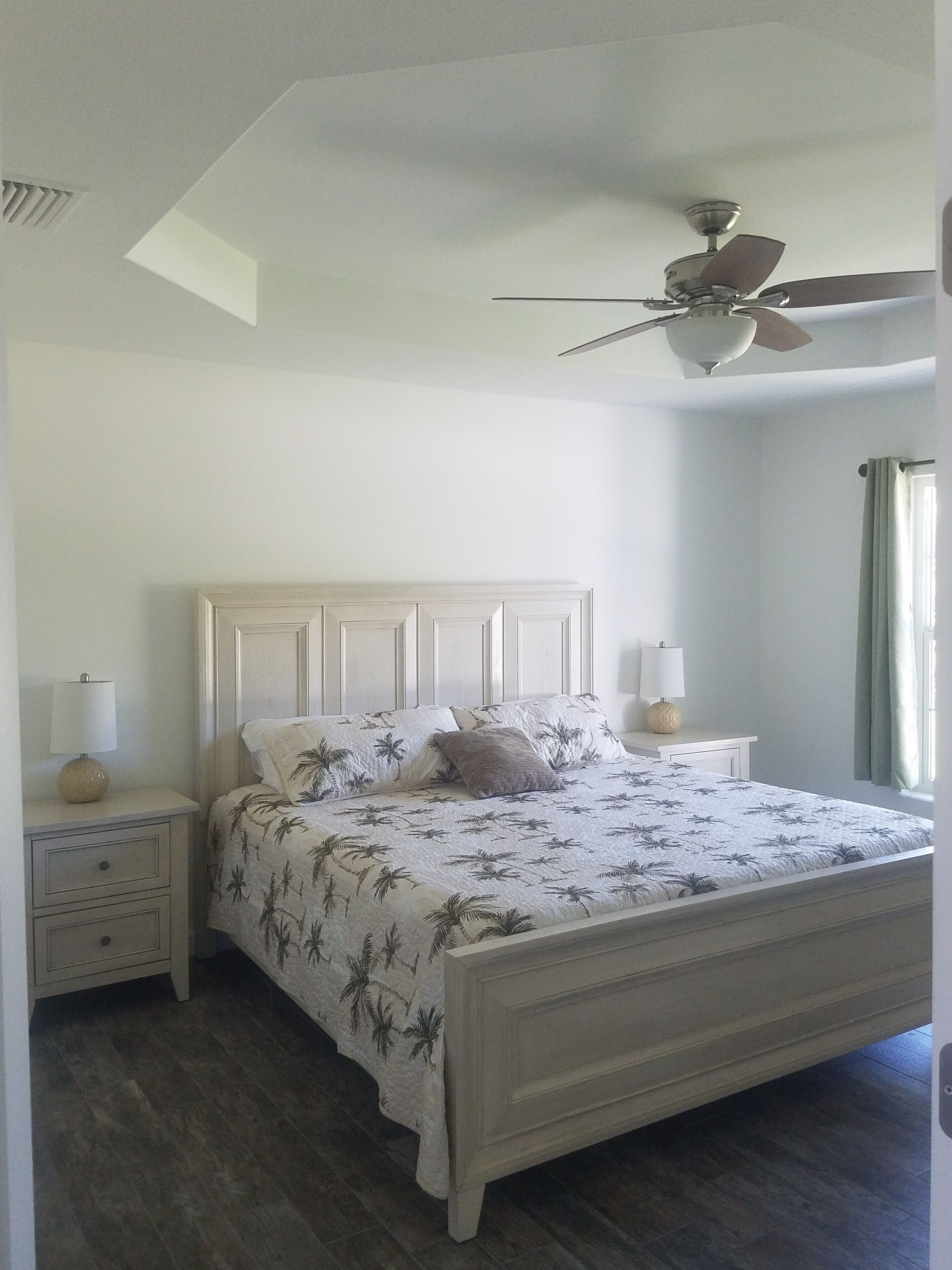 A bedroom with a large bed, two bedside tables with lamps, a ceiling fan, and a window with curtains.