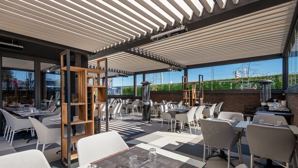 Un espacio al aire libre con mesas y sillas, una estantería de madera y vistas a la ciudad a través de amplios ventanales.