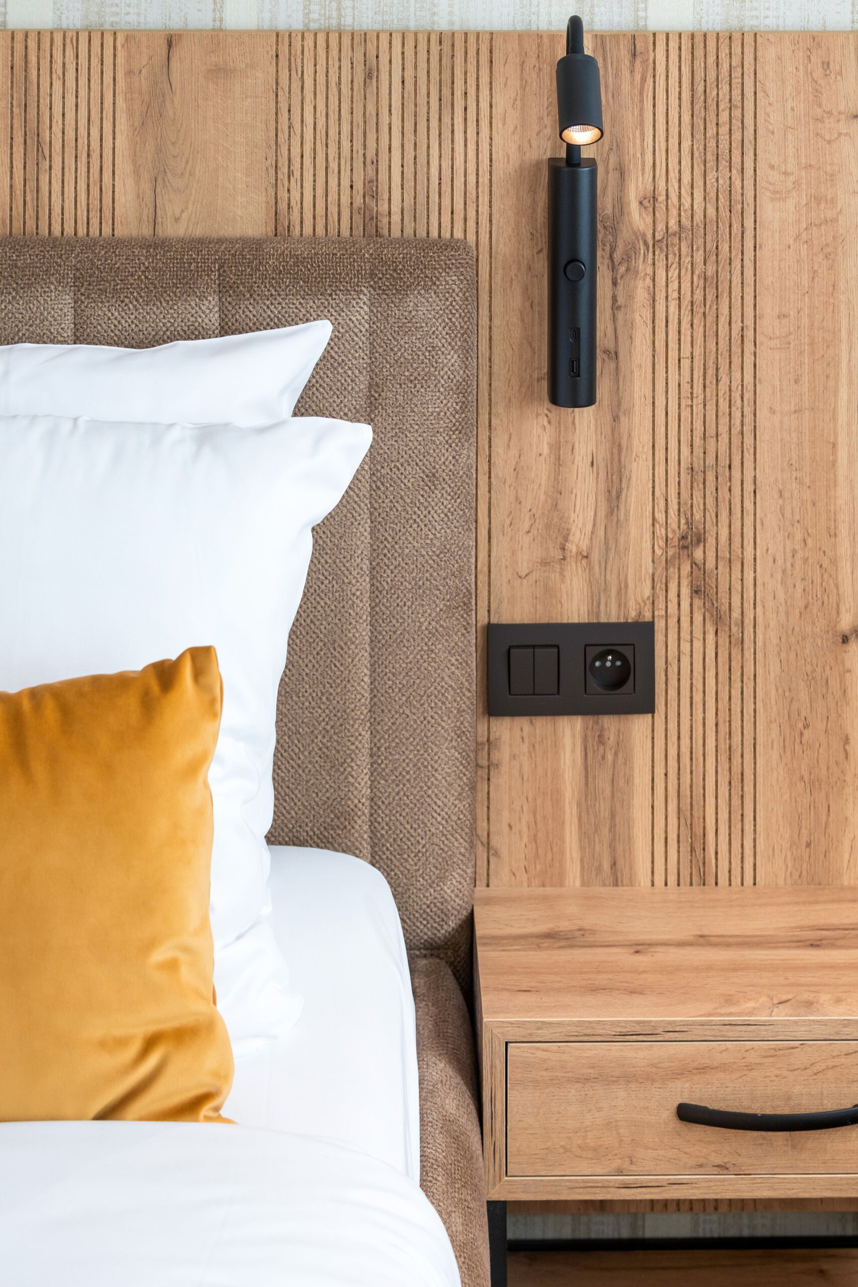 Una cama con almohadas blancas y mostaza, cabecera de madera, mesita de noche con cajón y aplique de pared.