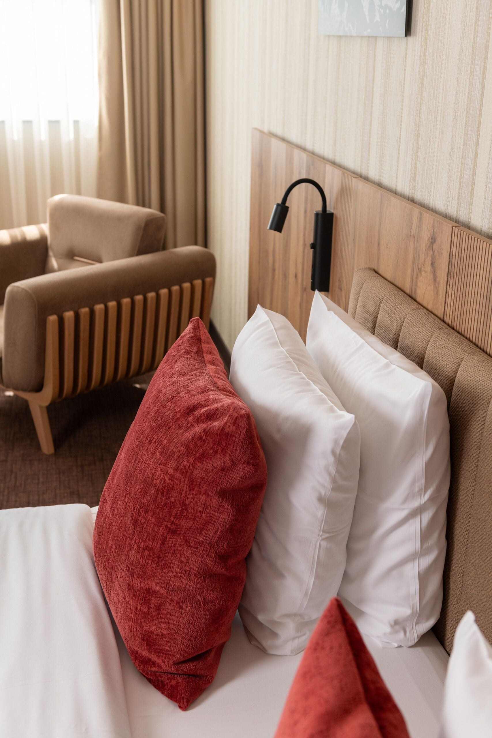 A hotel room with a bed, a red and white pillow, a wooden headboard, a bedside lamp, and a window with curtains.