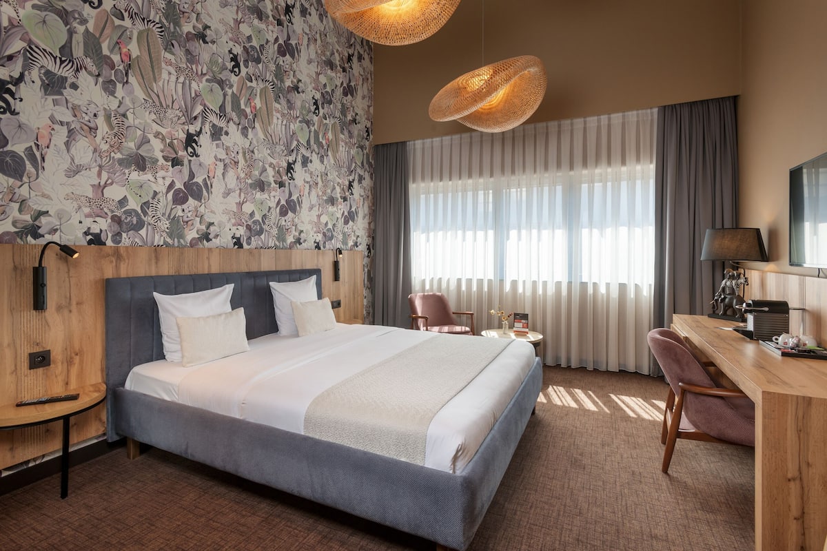 Une chambre d’hôtel avec un grand lit, un bureau, une chaise, une télévision et une fenêtre avec des rideaux.