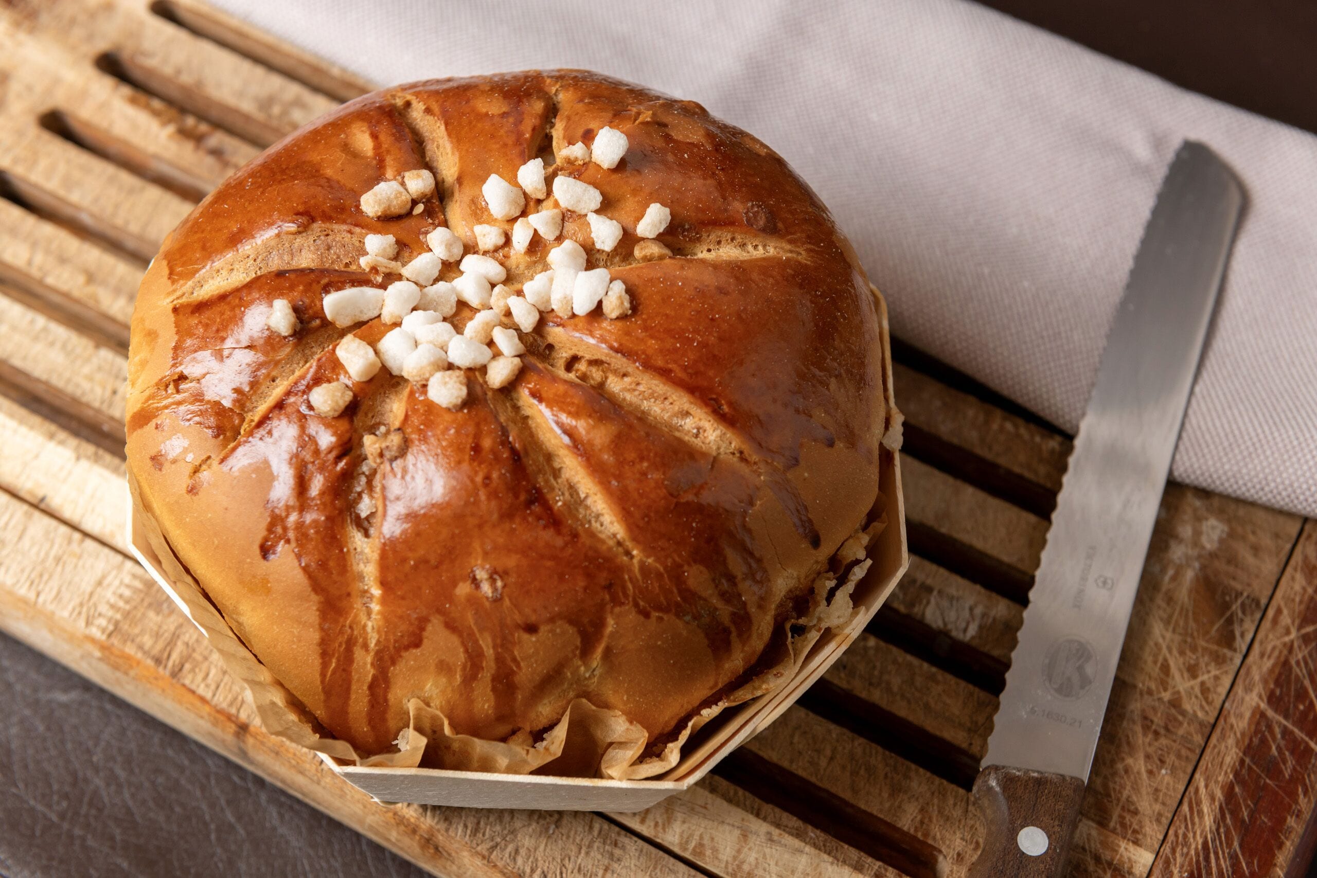 A loaf of bread with a crusty exterior and a glossy finish, placed on a wooden cutting board with a knife beside it.