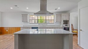A modern kitchen with a large island, white cabinetry, and a window providing a view of greenery outside. - TANGERINE HOUSE (Baton Rouge)