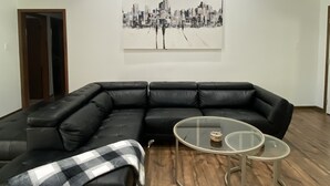 A spacious living room with a black sectional sofa, a glass coffee table, and a cityscape painting on the wall. - TANGERINE HOUSE (Baton Rouge)