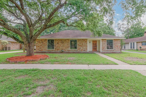 A single-story brick house with a front porch, a large tree in front, and a well-maintained lawn.