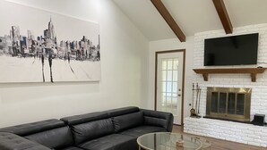 A modern living room with a black leather sectional sofa, a glass coffee table, and a fireplace with a TV above it. - TANGERINE HOUSE (Baton Rouge)