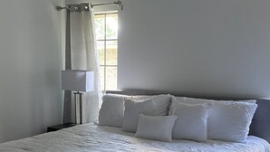 A neatly made bed with white linens, a bedside table with a lamp, and a window with curtains. - TANGERINE HOUSE (Baton Rouge)
